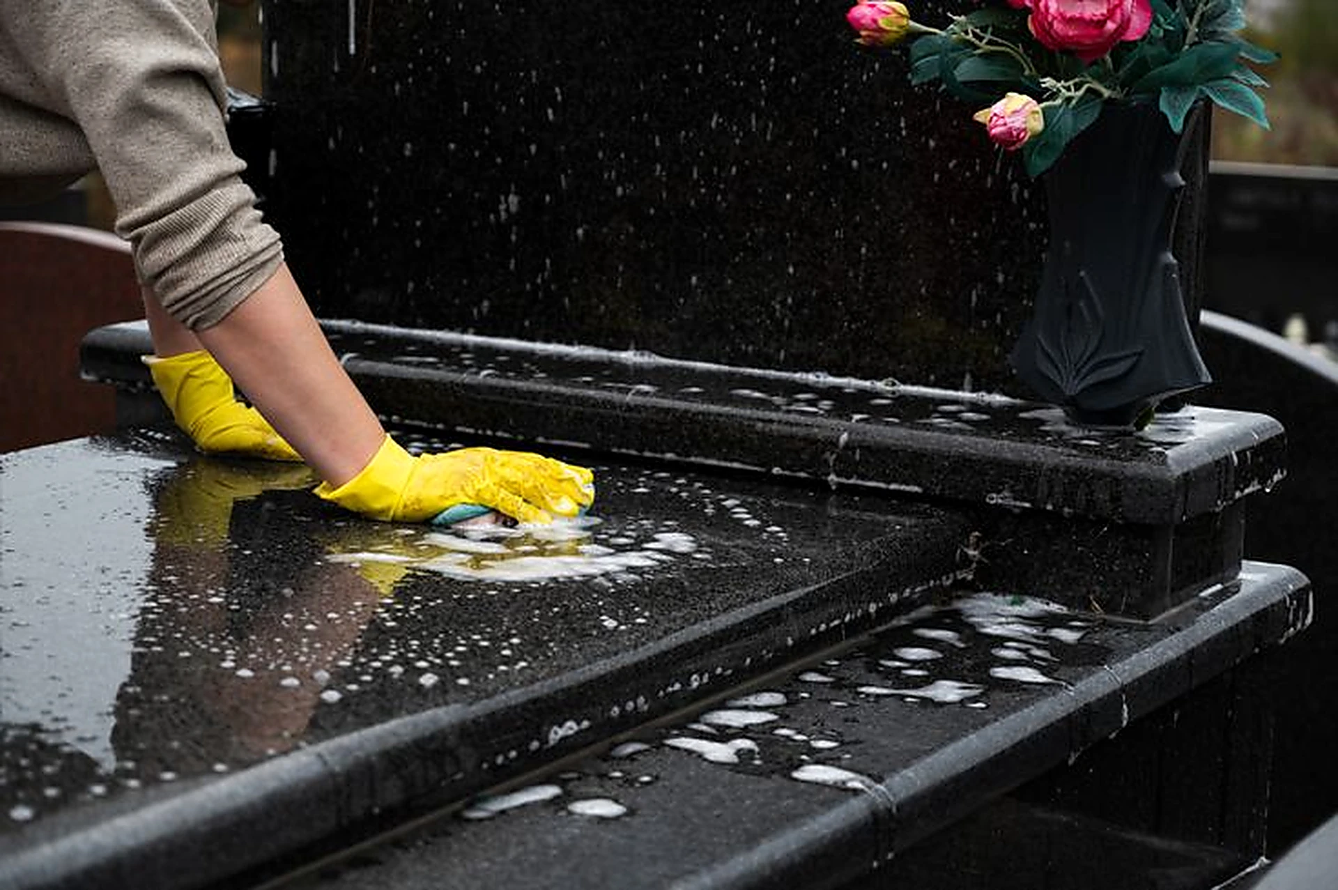 Gloved hands carefully washing a polished black granite headstone with soapy water